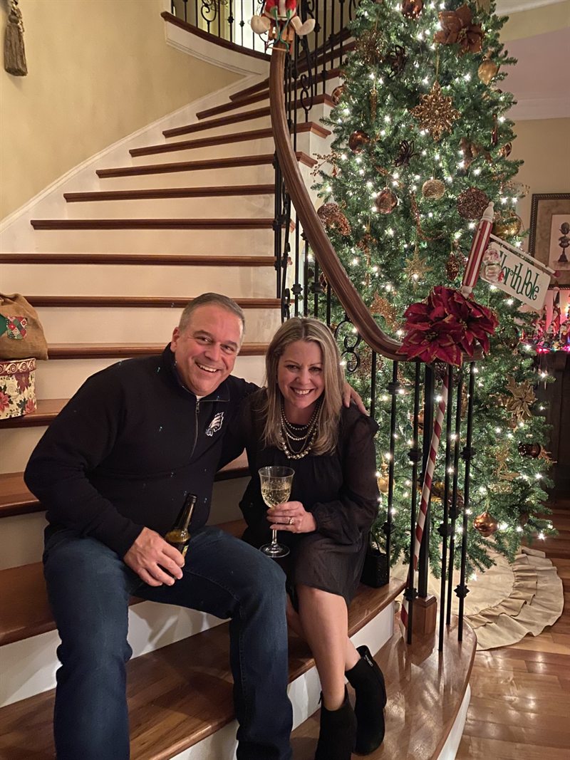 Chris and Kelley sitting on the staircase beside a lit Christmas tree, drinks in hand